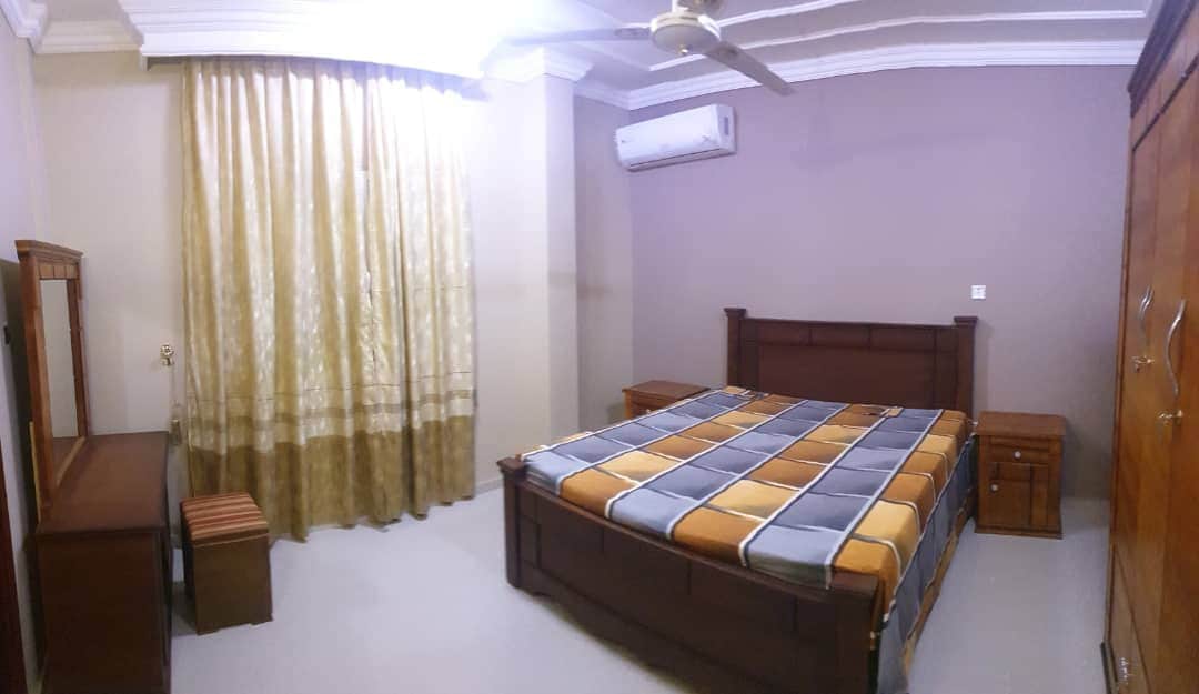 A spacious bedroom is furnished with a large wooden bed, featuring a multicolored blanket. Two nightstands are positioned on either side of the bed. A wooden dresser is placed near a window, adorned with light-filtering golden curtains. A ceiling fan is installed, and an air conditioning unit is visible.