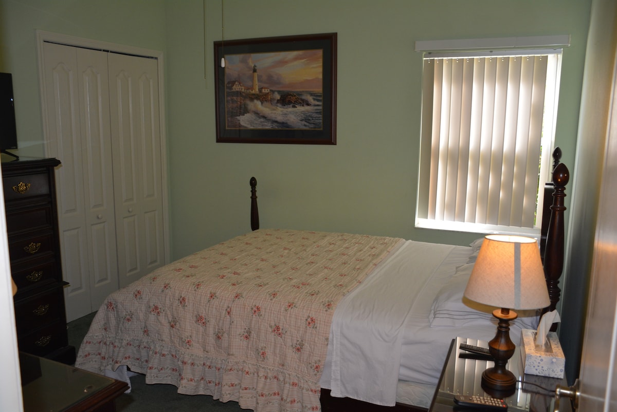 A serene bedroom is featured, with a wooden bed frame covered by a soft, floral-patterned quilt. A dresser stands against one wall, complemented by a lamp on a bedside table. Natural light filters through the blinds, enhancing the calming atmosphere.
