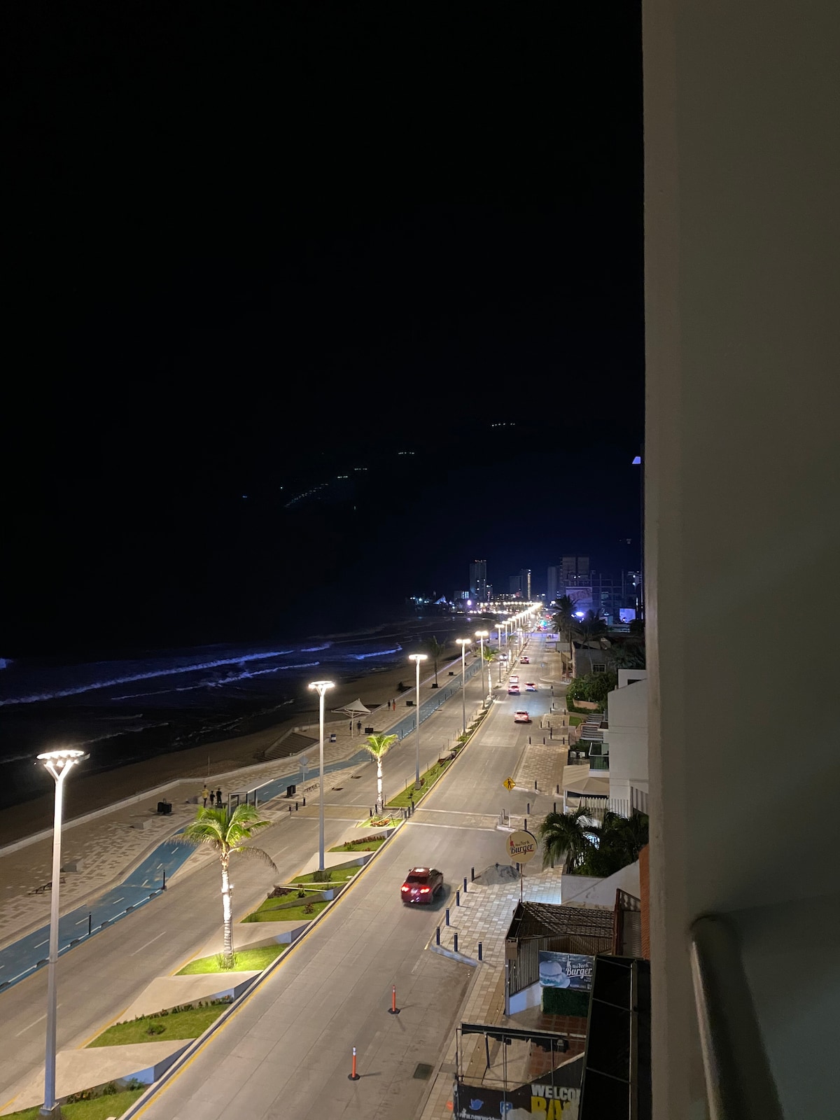 A nighttime view from the balcony captures the illuminated promenade lined with palm trees. The street is peaceful, featuring light posts that guide the way. In the distance, the ocean waves gently crash against the shore.