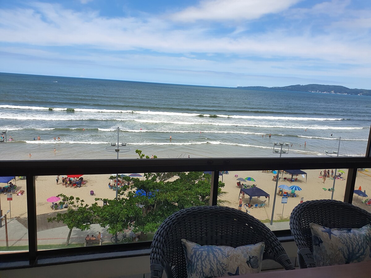 A spacious balcony offers a stunning view of the ocean and beach. Waves gently lap against the shore, where beach umbrellas and sunbathers are visible. Comfortable seating is positioned on the balcony, providing a perfect spot to enjoy the scenic surroundings.