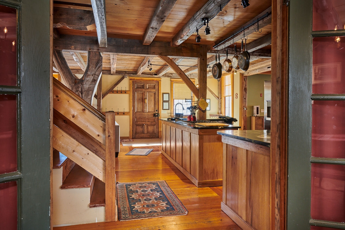 An open kitchen area features wooden cabinetry and a center island with seating. Exposed beams and warm wooden floors create an inviting atmosphere. A staircase is visible leading to the upper level, and a decorative rug adds a touch of color to the space.
