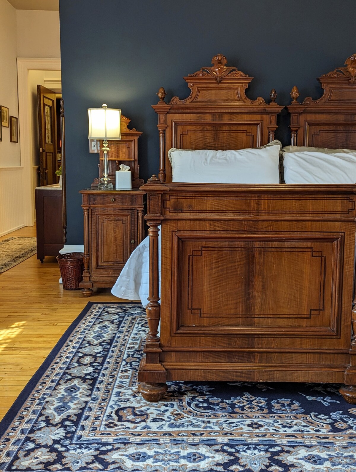 The room features a decorative wooden King-sized bed with intricate carvings and plush bedding. A bedside lamp provides soft illumination, complementing the dark accent wall. An elegant area rug with a floral pattern enhances the warm wooden flooring.