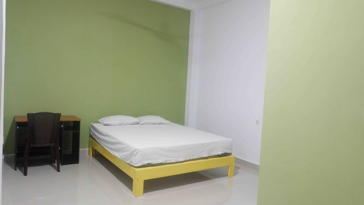 A bedroom features a spacious bed with a light-colored mattress resting on a yellow frame. A desk with a chair is positioned against the wall, and a vibrant green accent wall complements the light-colored floor and surrounding neutral walls.