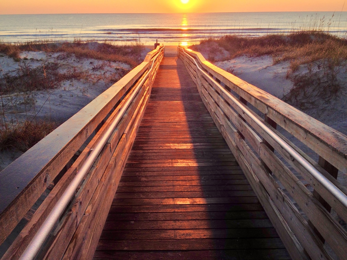 A wooden boardwalk guides the way toward the ocean at sunset. Soft golden light reflects off the sandy beach and water, while tall grasses frame the path on either side. The peaceful scene emphasizes a serene transition from land to sea.