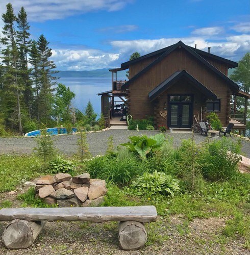 A newly constructed chalet with a wooden exterior is framed by lush greenery, featuring a spacious patio and a view of the water. A fire pit made of stones complements the outdoor area, while distant trees and clouds add to the serene landscape.