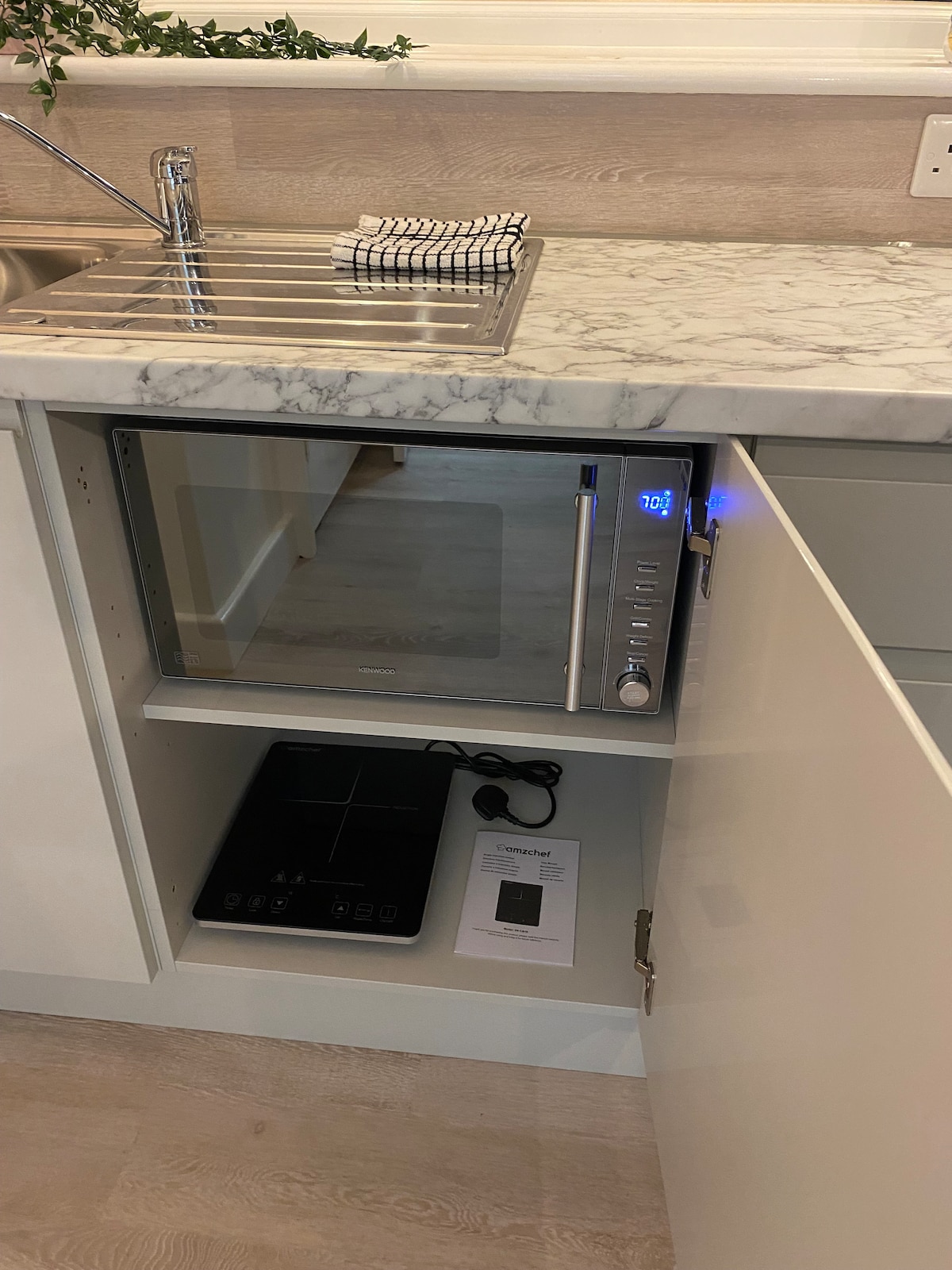 A modern kitchen area features a marble countertop with a sleek sink and a neatly positioned dish drying rack. Below, a microwave and a portable induction hob are housed in a cupboard, accompanied by an instruction manual, promoting a clean and functional space.