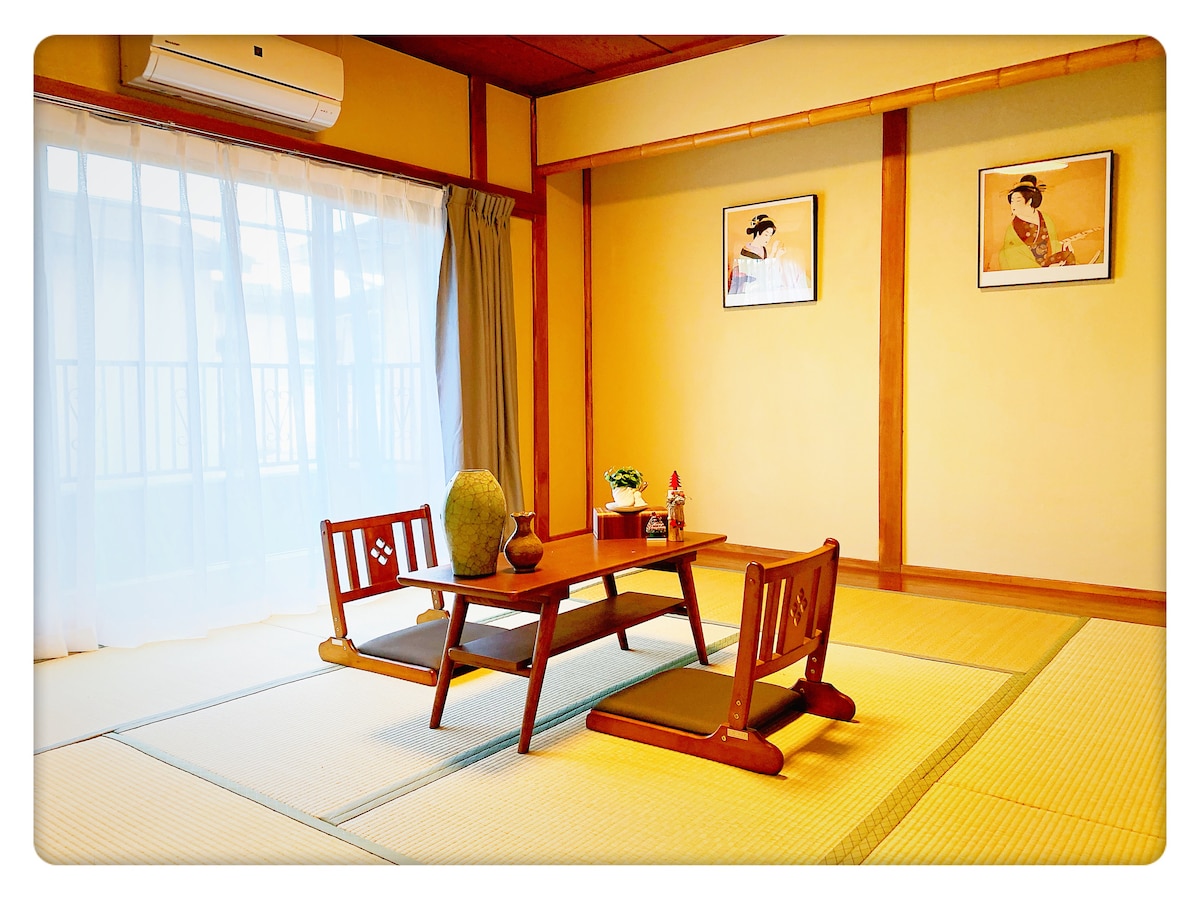 A traditional Japanese room features a low wooden table surrounded by two seating cushions. Sunlight filters through sheer curtains, illuminating the tatami mats. Two framed art pieces hang on the walls, adding cultural charm to the serene space.