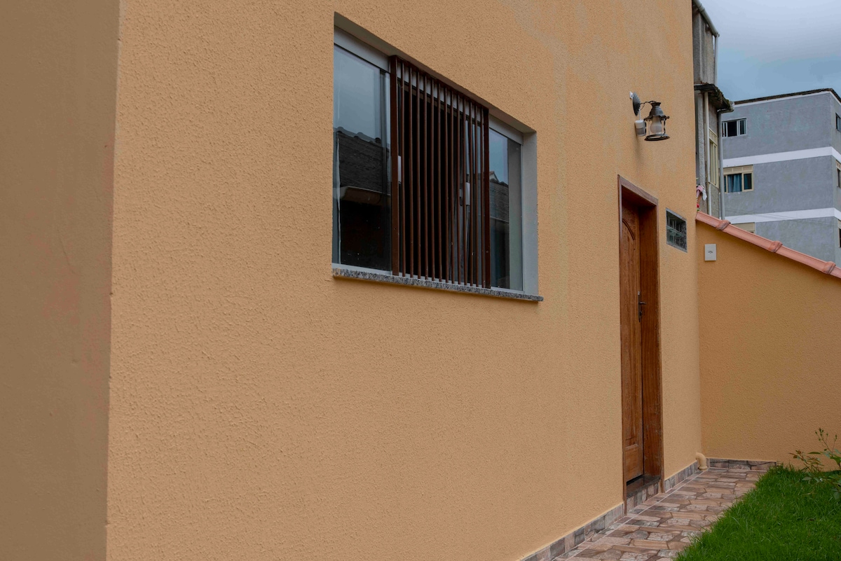 The exterior of the building features a smooth, light-colored wall with a large window partially covered by horizontal bars. A wooden door with simple hardware provides entry, while paved stones surround the entrance, maintaining a neat appearance. The area is well-lit and open.