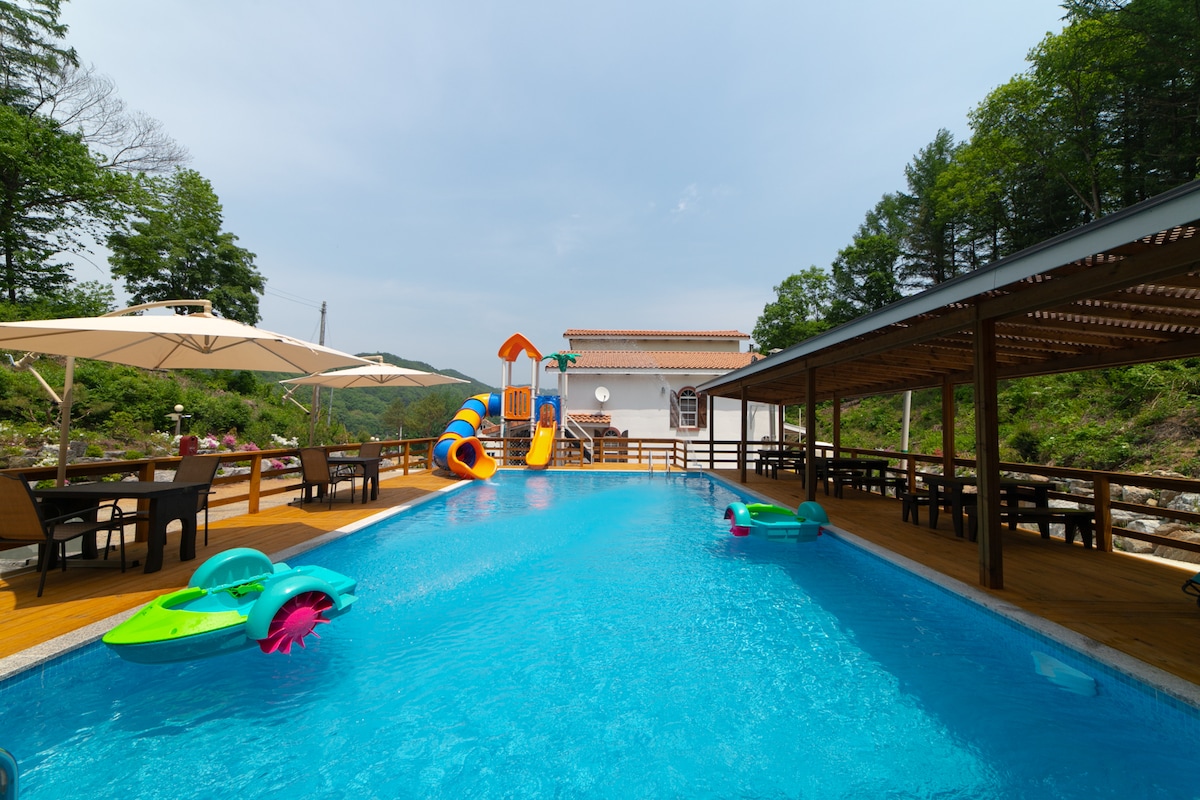 A refreshing outdoor swimming pool is depicted, featuring a colorful water slide. Lounge chairs and shaded areas surround the pool, providing ample space for relaxation. Two floating chairs are visible in the water, while a building is present in the background among lush greenery.