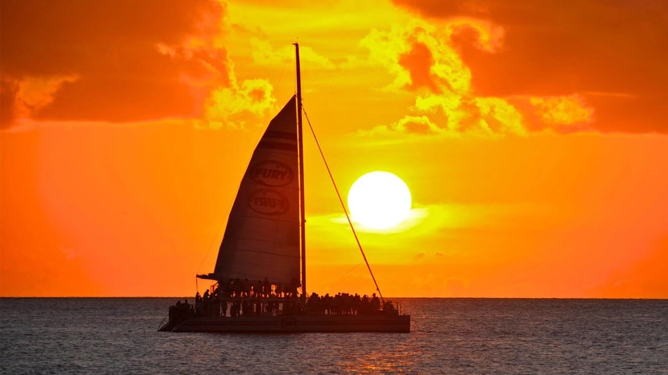 A sailing catamaran is silhouetted against a vibrant orange sunset over the ocean. The sun, large and bright, is positioned just above the horizon, surrounded by warm hues and soft clouds.