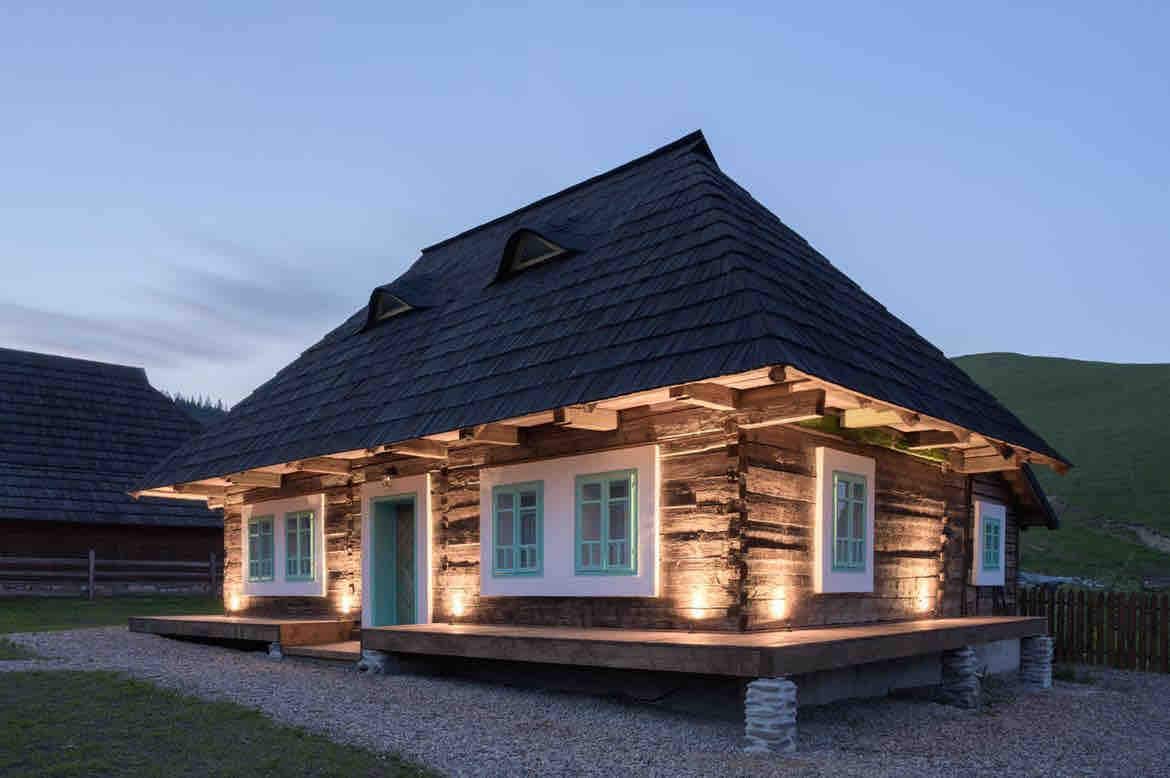 The traditional wooden house features a dark, steeply pitched roof and is illuminated by soft outdoor lighting. The structure showcases exposed wood beams and large, pale blue windows, creating a harmonious blend with the natural surroundings.