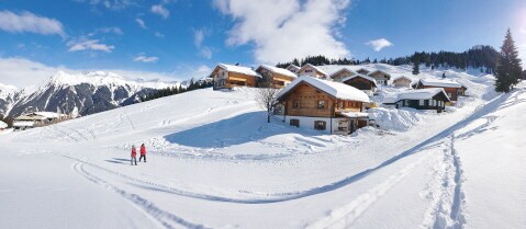 A serene winter landscape features several wooden chalets blanketed in fresh snow, surrounded by snow-covered mountains. A clear blue sky complements the scene, while two individuals can be seen walking along a snowy path, highlighting the peaceful environment.