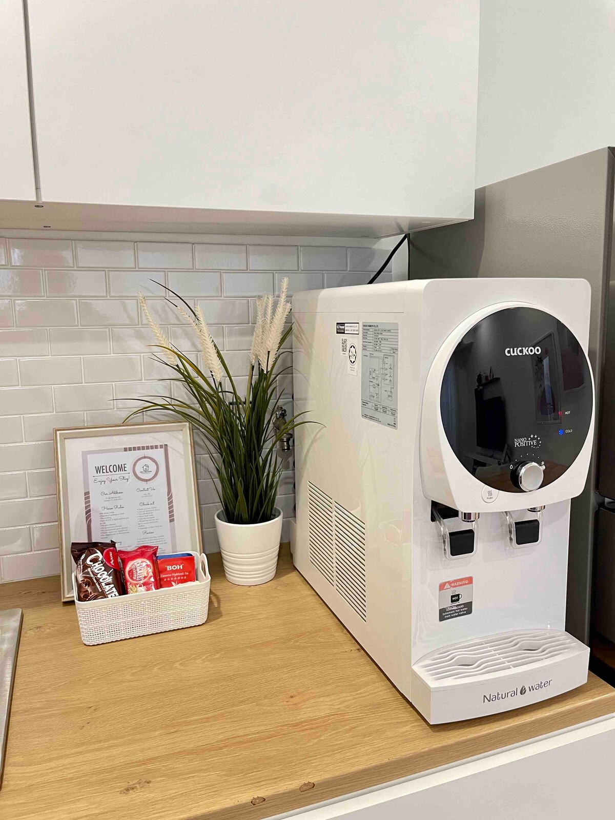 A modern water dispenser is positioned on a wooden countertop, featuring two taps for hot and cold water. Beside it, a woven basket contains a selection of instant beverages. A framed welcome sign and a potted plant add a touch of warmth to the space.