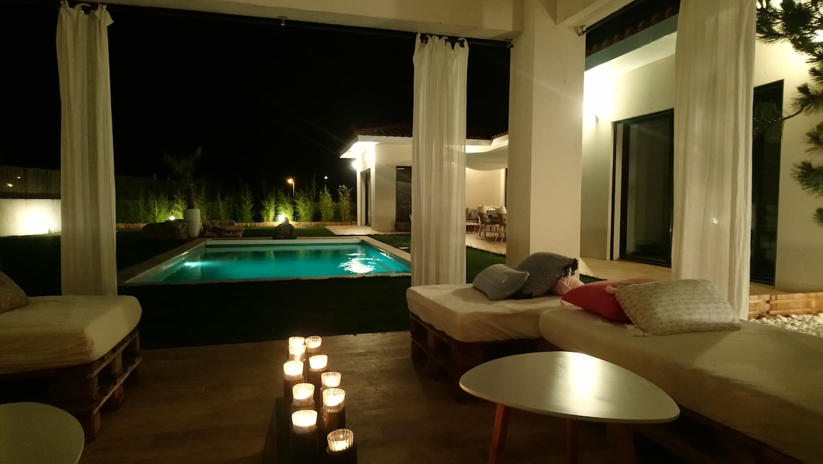 A serene outdoor space is illuminated softly by candlelight, with comfortable loungers positioned nearby. A private pool is visible in the background, surrounded by grass and landscaped greenery. Sheer curtains frame the area, creating a quiet retreat for relaxation.
