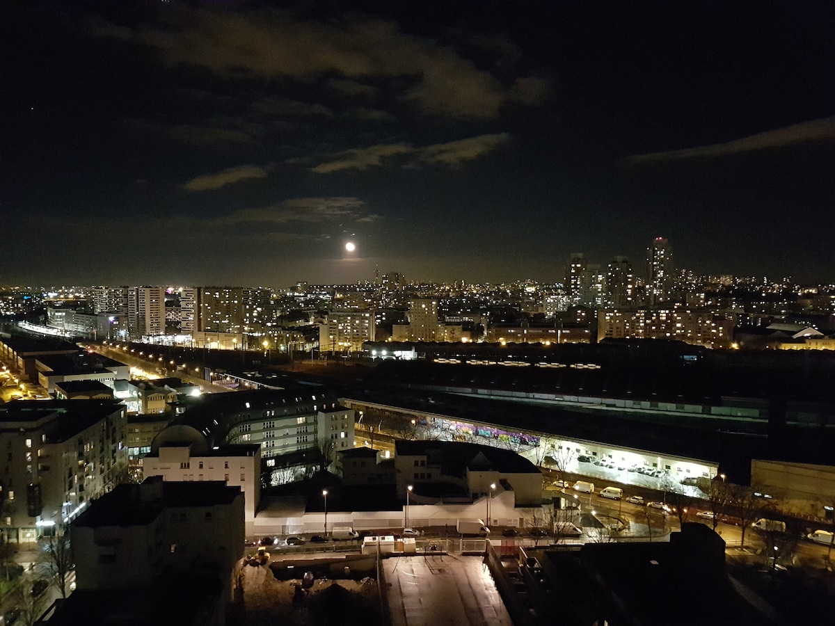 A panoramic view captures the city skyline at night, illuminated by streetlights and the glow of the moon. Distant buildings reflect a warm light, while a winding train track runs through the foreground, adding depth to the urban landscape.