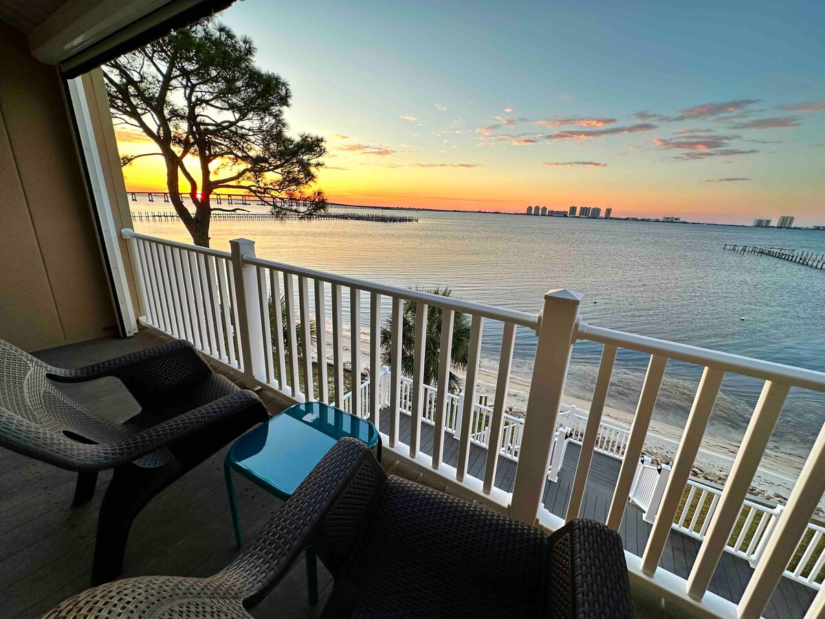 A private balcony is shown, featuring two chairs and a small table overlooking the water. The scene captures a vibrant sunset reflecting on the calm surface of Santa Rosa Sound, with distant buildings visible along the horizon.