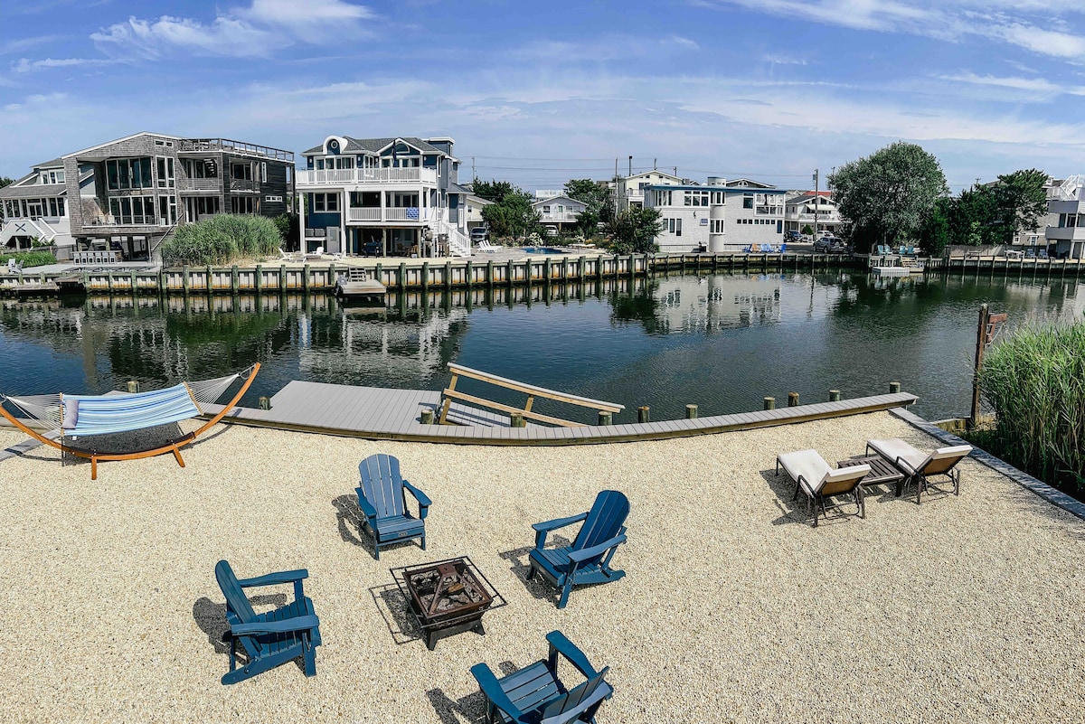 The waterfront area features a selection of blue Adirondack chairs arranged around a firepit, with a sandy surface leading to the water's edge. A hammock is positioned nearby, and lounge chairs are scattered for sunbathing, all surrounded by greenery and a calm canal view.