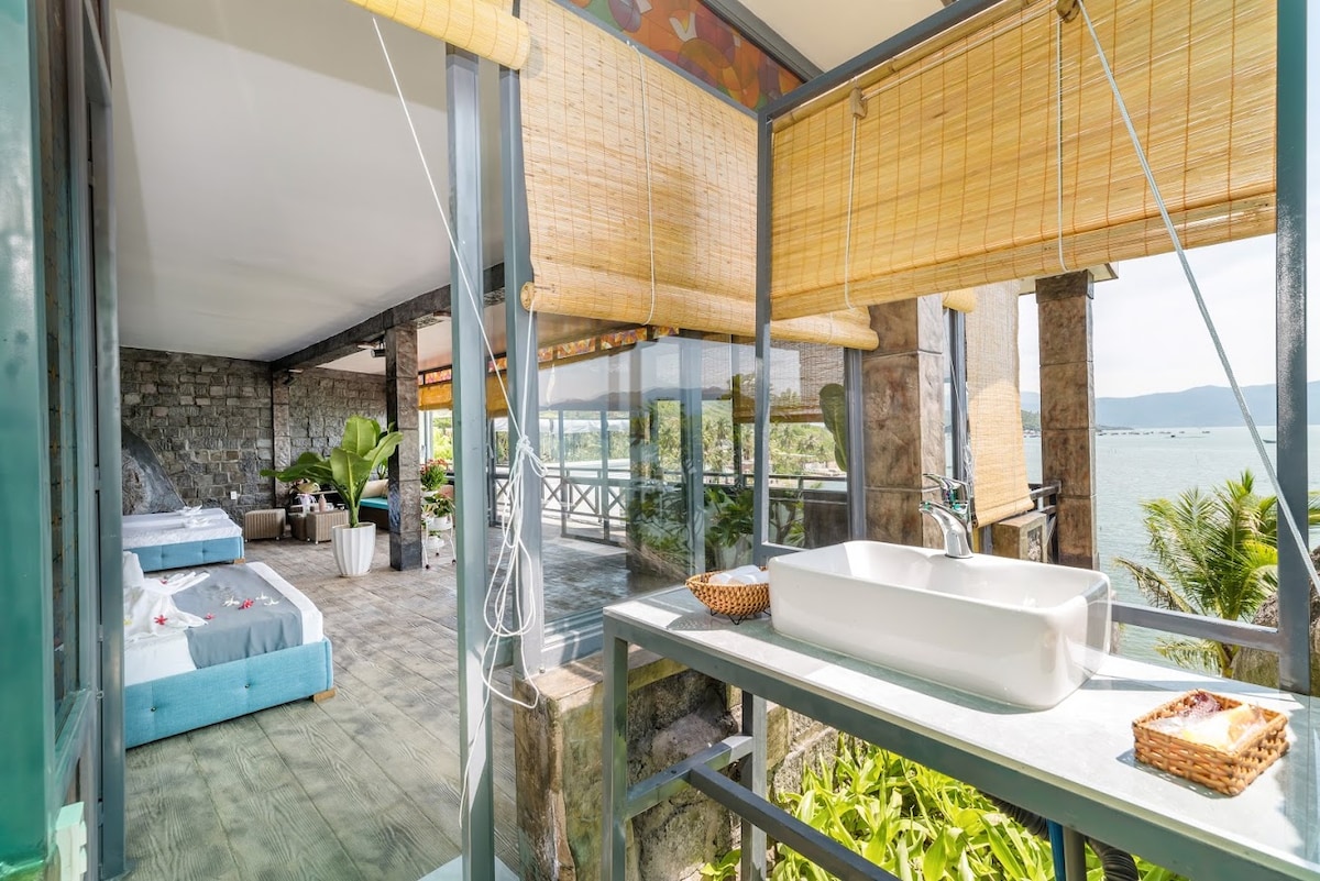 An open space is illuminated by natural light, showcasing a blend of indoor and outdoor living. A stylish vanity with a modern sink is situated beside a large window, offering views of the ocean and surrounding greenery. Comfortable seating areas invite relaxation.