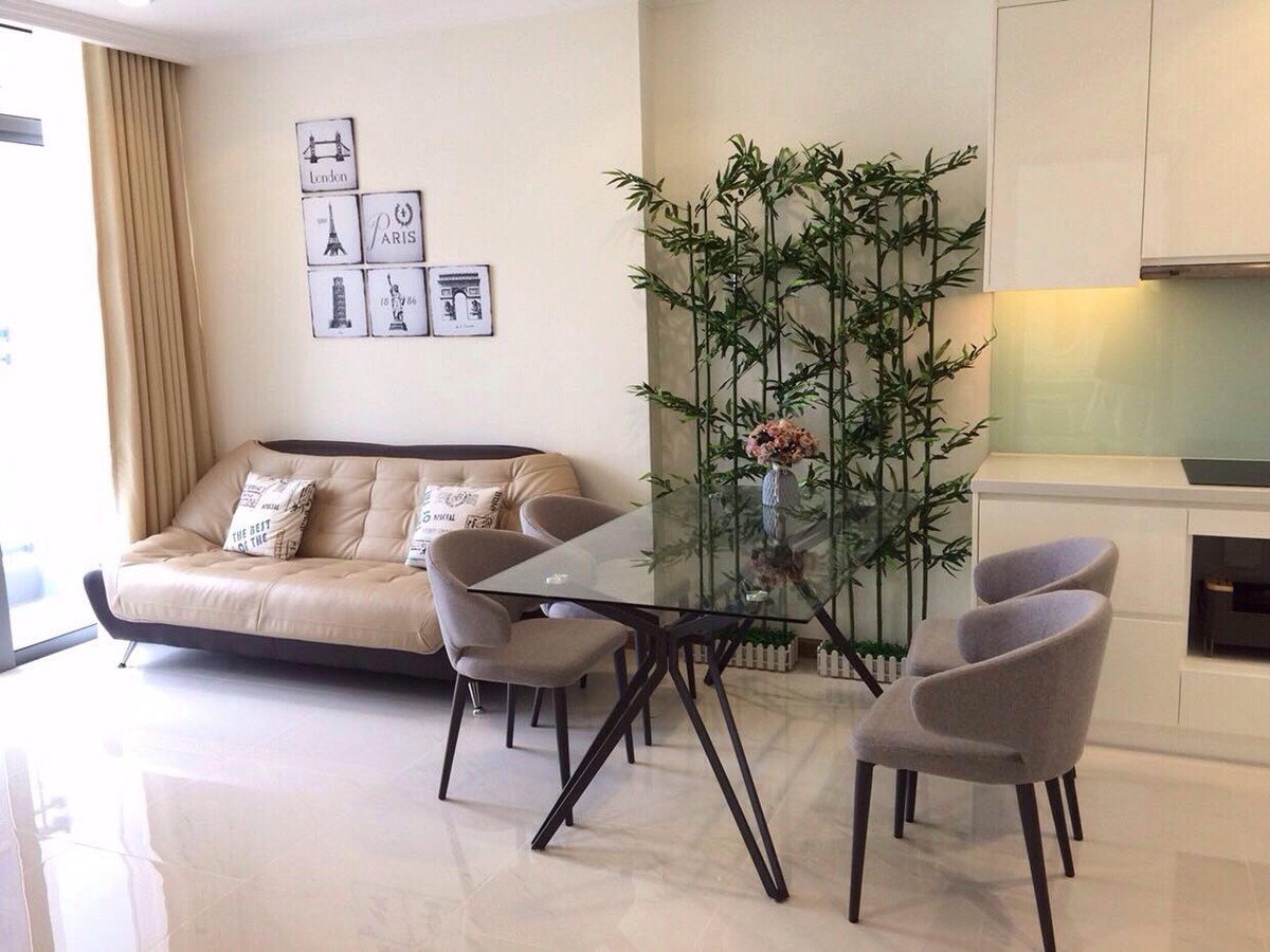 A modern living space features a light-colored sofa accompanied by decorative cushions. A sleek glass dining table is surrounded by four upholstered chairs. A decorative plant adds greenery, while artworks are displayed on the wall, enhancing the contemporary ambiance.