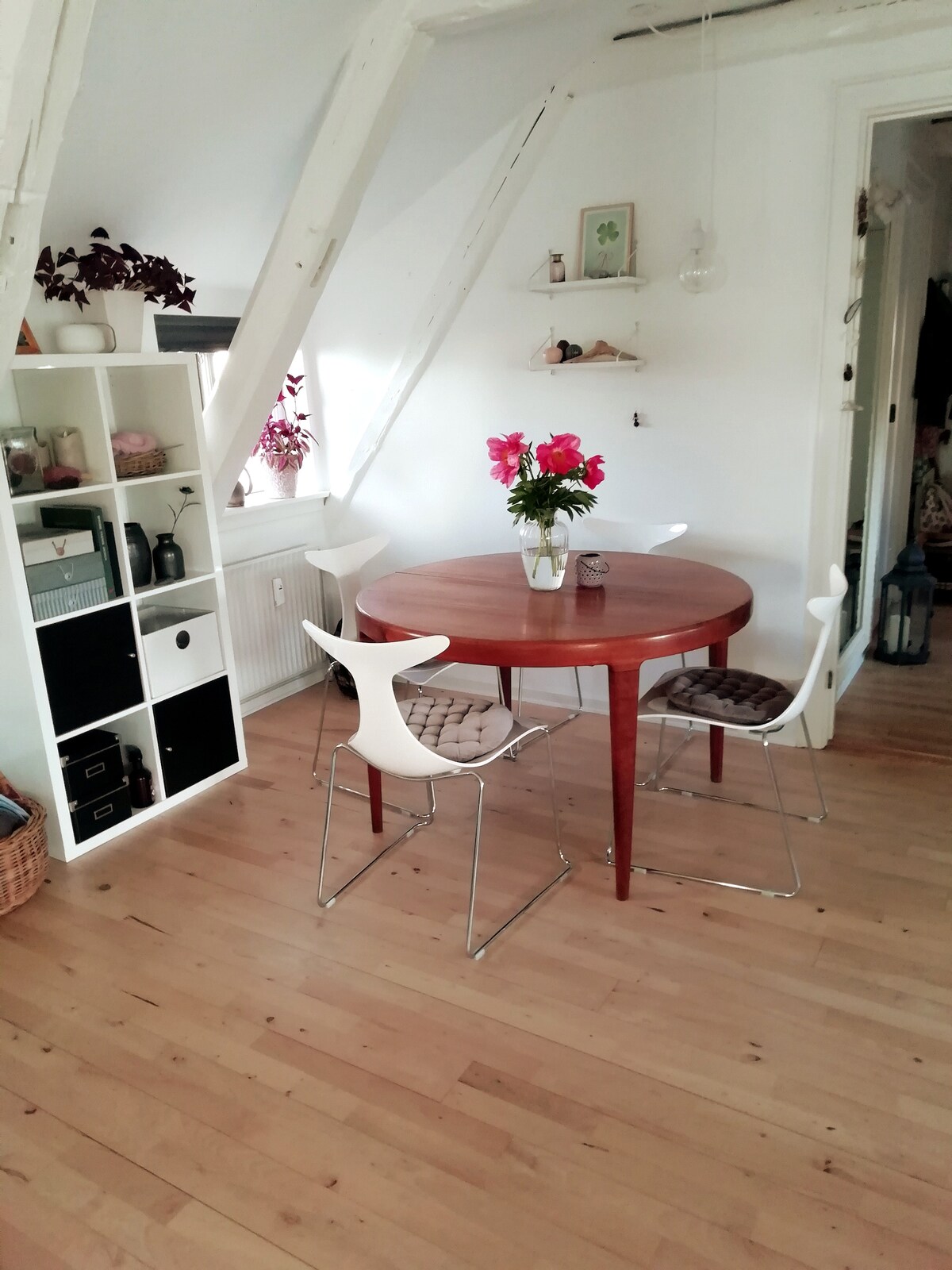 A round wooden dining table is positioned near a window, surrounded by three modern chairs. A vase of flowers serves as a centerpiece. A shelf with decorative items is seen in one corner, complemented by wooden flooring that enhances the light atmosphere.