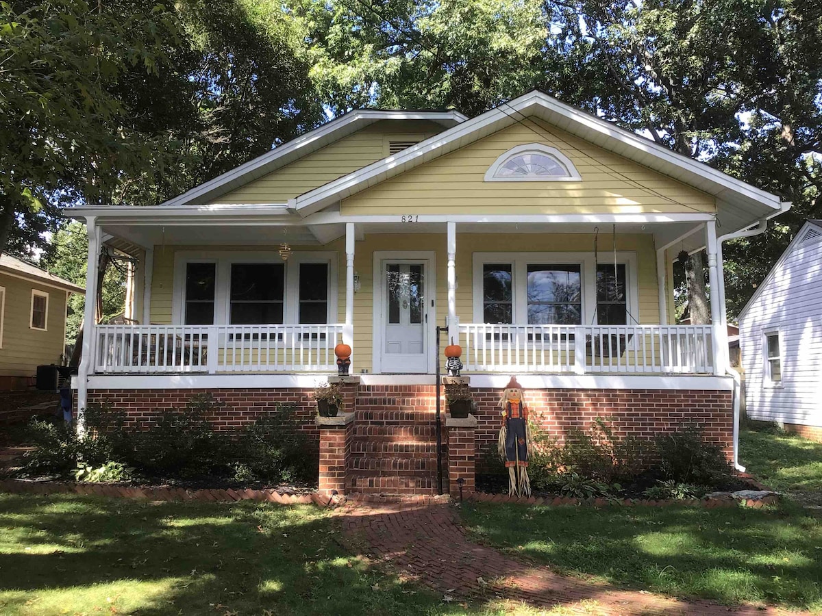 A charming yellow house features a spacious front porch framed by white railings. Brick steps lead to the entrance, while large windows invite natural light. The landscaped yard includes colorful seasonal decorations, enhancing the welcoming appearance of the home.