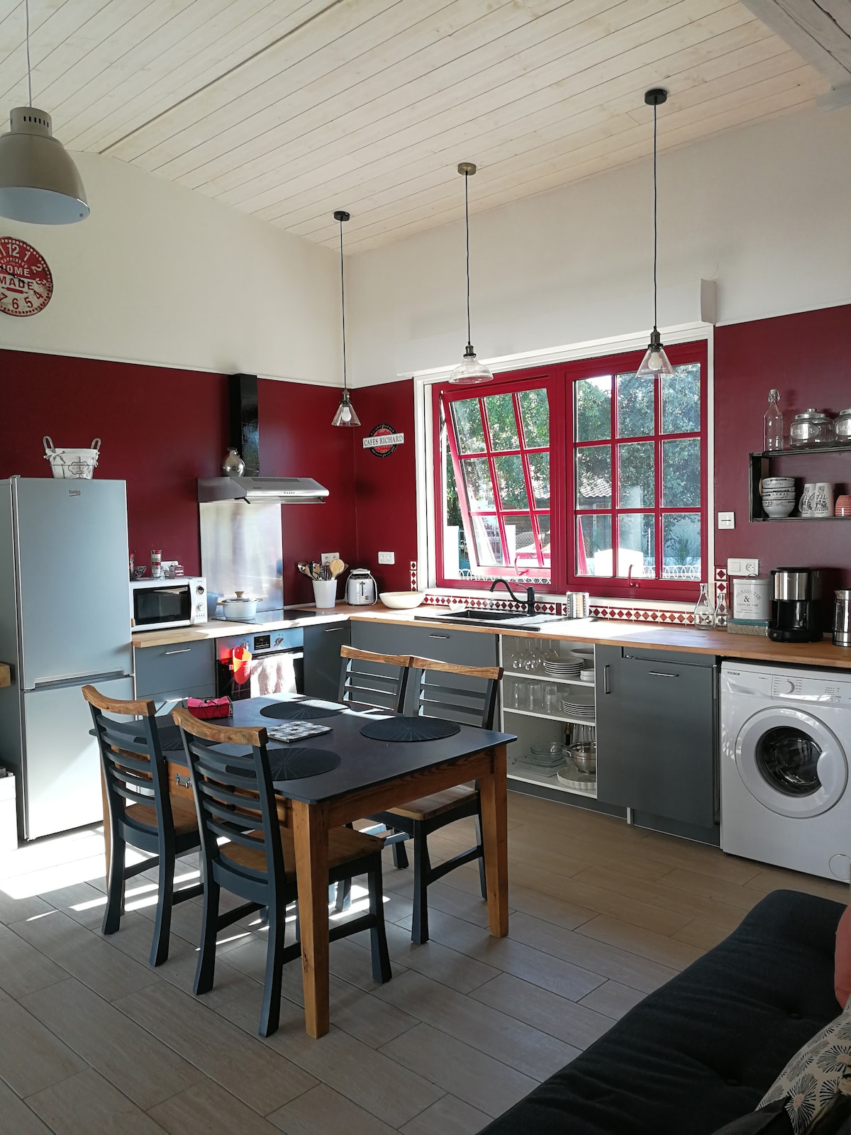 A modern kitchen features red accent walls complemented by white cabinetry. Stainless steel appliances include a refrigerator, microwave, and dishwasher. A wooden dining table with four black chairs is situated near large windows, allowing natural light to brighten the space. A washing machine is visible to the right.
