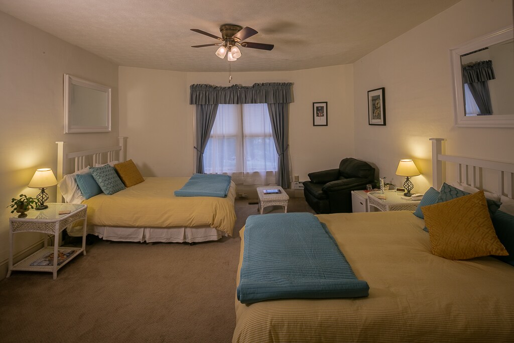 The spacious room contains two queen-size beds, each with minimalist white frames and soft yellow bedding. A cozy seating area features a dark-colored chair beside a small table. Natural light is filtered through a large window with light blue curtains.