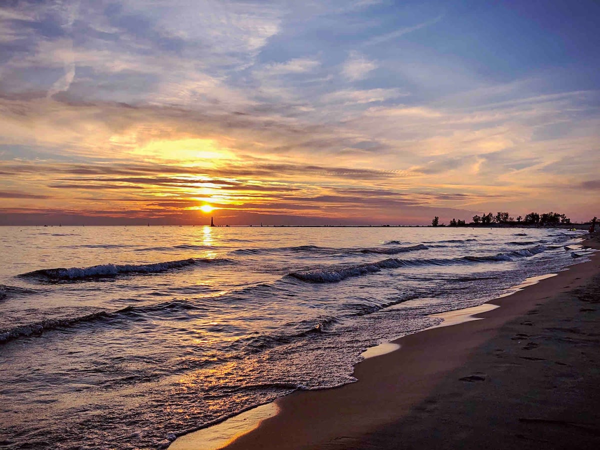 A serene view of a beach at sunset showcases gentle waves lapping against the shore. The sky is filled with soft hues of orange, pink, and purple, reflecting on the water's surface. Silhouetted trees line the distant beach, adding to the peaceful ambiance.