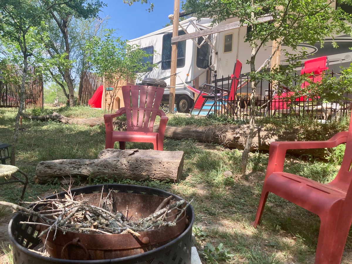 A cozy outdoor area is shown with a fire pit surrounded by several red chairs. The RV is partially visible in the background, nestled among trees. Sunlight filters through the leaves, highlighting the inviting space for relaxation and outdoor gatherings.