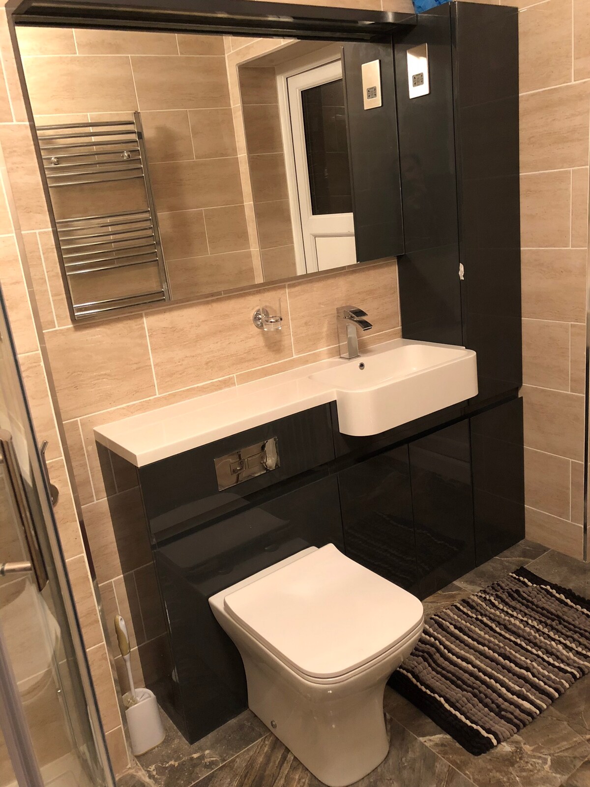 The bathroom features contemporary fittings, including a sleek vanity with an integrated sink and a mirror above. Soft beige tiles line the walls, complemented by a dark floor. A glass shower is visible, and a towel rack adds functionality. A striped mat is placed in front of the toilet.