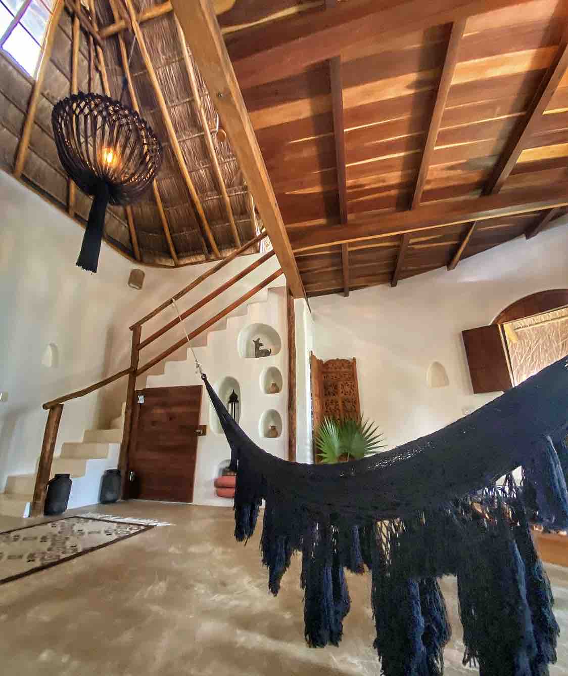 A cozy living area features a dark-hued hammock suspended from the ceiling, inviting relaxation. Natural wood beams and a woven chandelier create a rustic atmosphere, while light reflects off the smooth walls, highlighting niches displaying decorative elements. A staircase leads to an upper level.