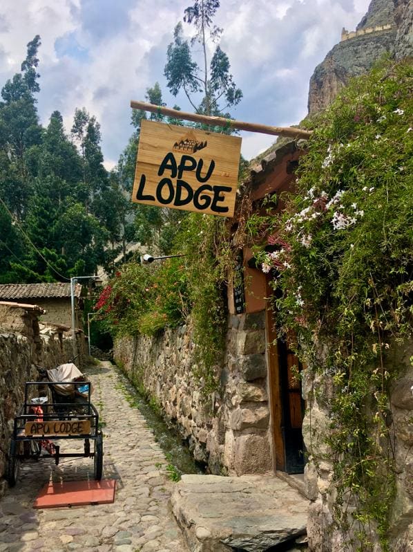 A rustic stone pathway leads to the entrance of Apu Lodge, marked by a wooden sign displaying the name. Lush greenery and colorful flowers surround the stone walls, creating a welcoming atmosphere along the pedestrian path.