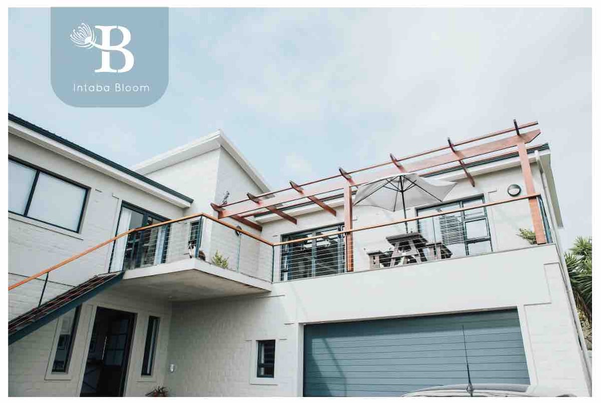 The apartment entrance is situated above a garage, featuring a balcony with a wooden pergola and an outdoor seating area. A table with chairs and an umbrella provides a shaded space, while the exterior is characterized by clean lines and a modern design.