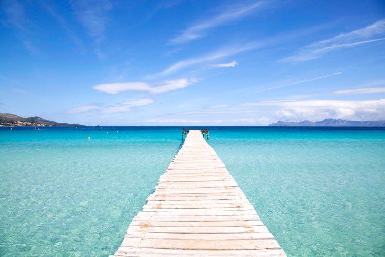 A wooden pier extends into the clear turquoise waters, creating a path toward the horizon. Waves gently lap against the sides, and the sky above is bright with scattered clouds, enhancing the serene atmosphere of the coastal scene.