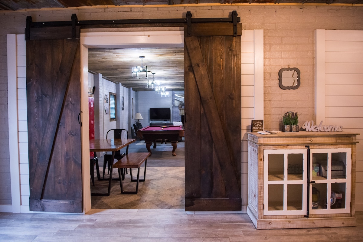 A spacious entryway is framed by large wooden barn doors. Inside, an open living area features a pool table and comfortable seating arranged around a television. A wooden dining table sits nearby, adorned with simple decor that welcomes guests.