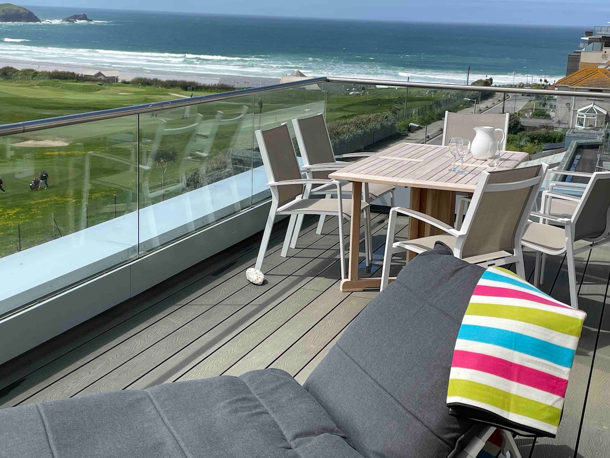 A spacious balcony features a light wood dining table surrounded by six chairs. A comfortable gray lounge chair is positioned nearby, adorned with a colorful striped throw. The expansive views encompass the golf course and Fistral Beach beyond, creating a serene outdoor space.