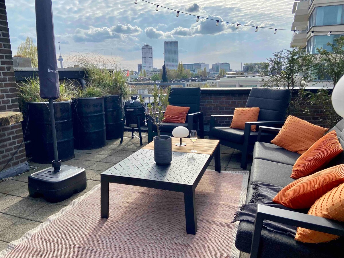 The spacious rooftop terrace offers stunning city views, featuring a lounge set with orange pillows arranged around a central table. Potted plants and a BBQ grill are visible, along with string lights overhead, creating a relaxed outdoor atmosphere.