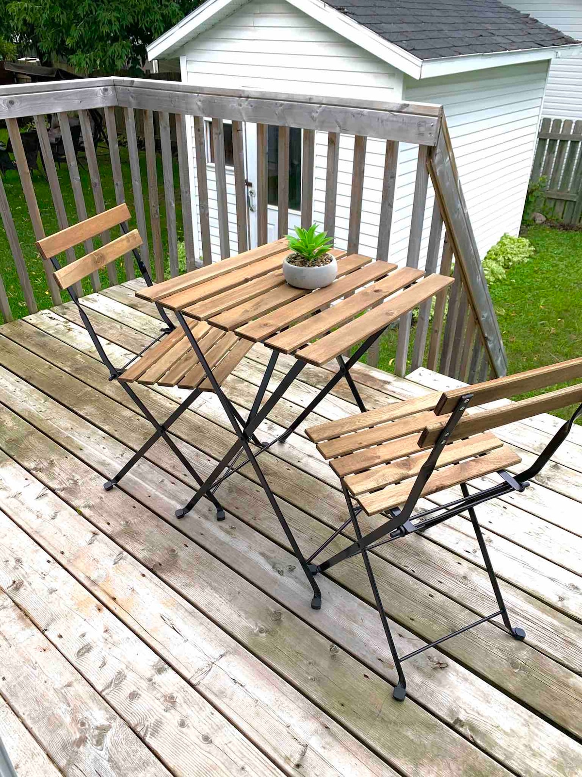 A small wooden table with matching foldable chairs is situated on a wooden deck. A potted plant rests on the table, with a view of a grassed area and a white shed in the background. The space offers a quiet outdoor setting.