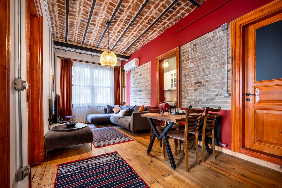 A spacious living room features exposed brick walls and a stylish wooden ceiling with beams. Comfortable seating is arranged around a coffee table, complemented by a dining area with wooden chairs. Warm lighting and rich colors create a cozy atmosphere, with large windows allowing natural light.