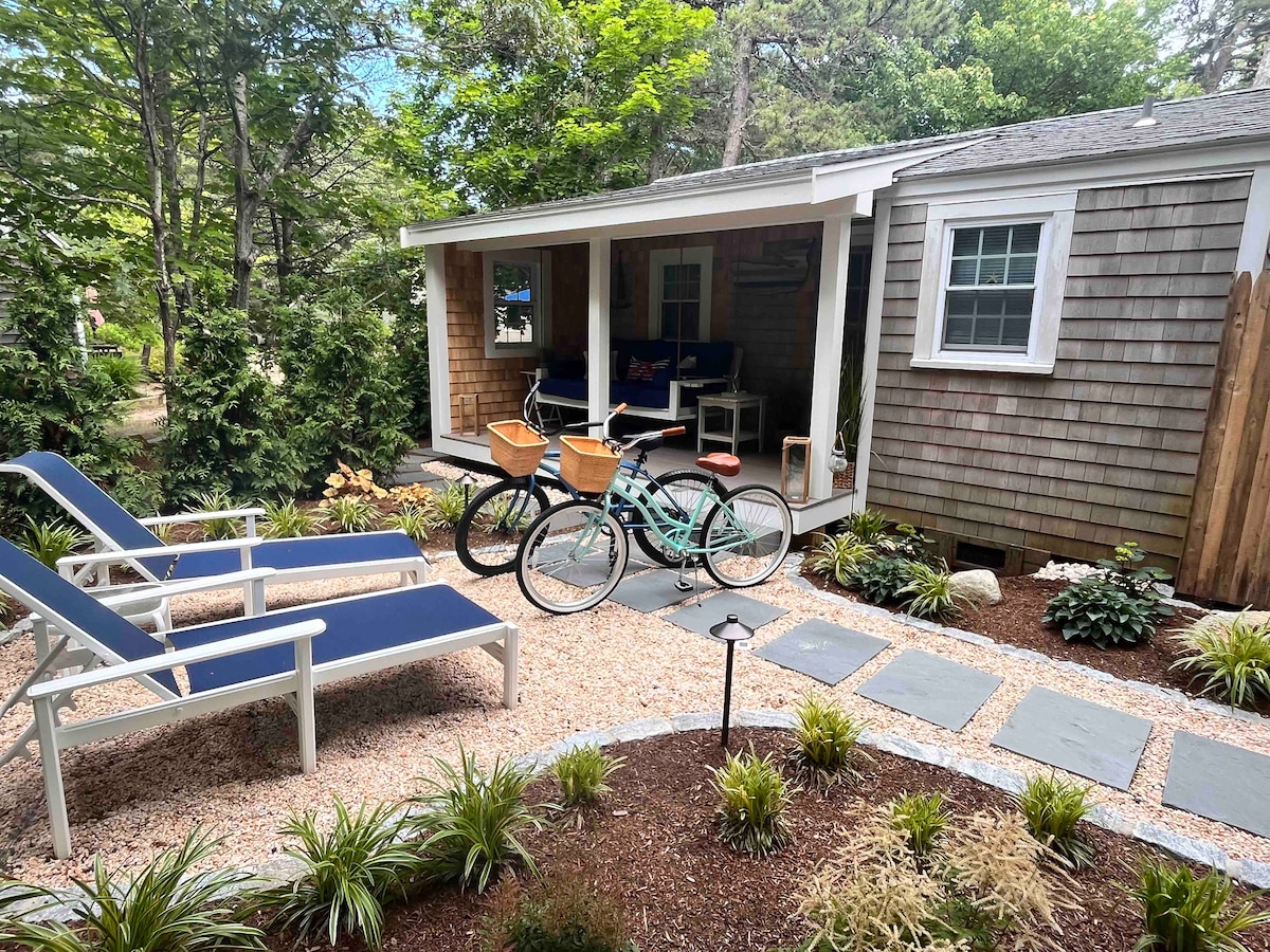 A charming outdoor area showcases two bicycles parked on a stone path, framed by lush greenery and a well-manicured garden. The cottage exterior features wooden shingles and a welcoming porch with seating, complemented by two lounge chairs positioned on the crushed stone patio.