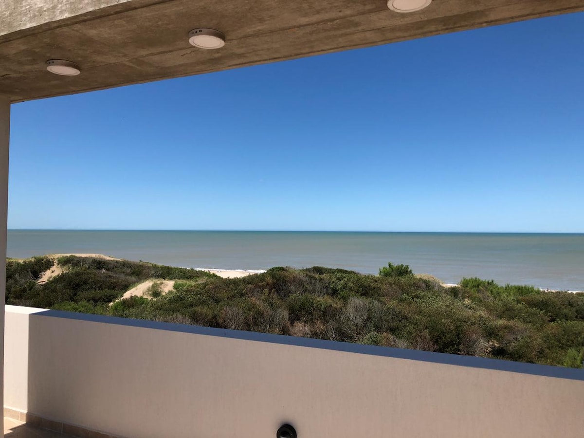 A wide view captures the horizon where the tranquil ocean meets the clear blue sky. Lush greenery lines the foreground, creating a natural border. The image is framed by a solid balcony railing, emphasizing the serene coastal setting.