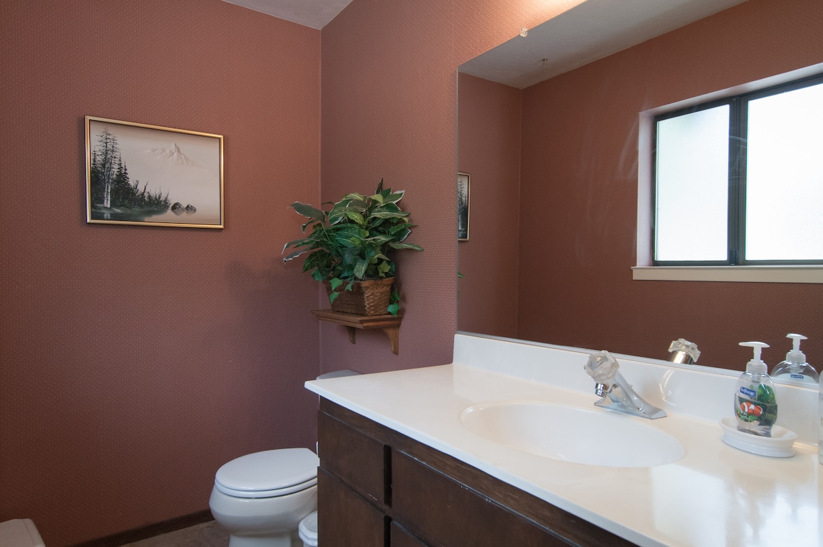 A private bathroom is equipped with a white countertop and a single sink. A dark wooden cabinet provides storage, and a large mirror is mounted above the sink. A window allows natural light, while a decorative plant adds a touch of greenery to the space.