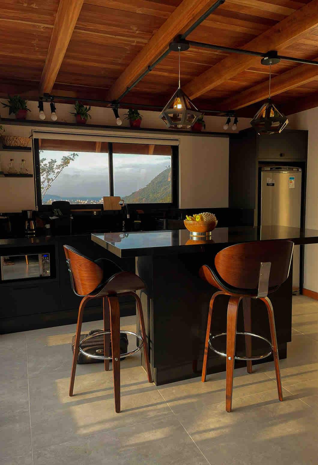 A modern kitchen features a sleek black countertop and matching bar stools. Large windows allow natural light to flood the space, showcasing a view of the surrounding landscape. Pendant lights hang from a wooden ceiling, creating an open and inviting atmosphere.