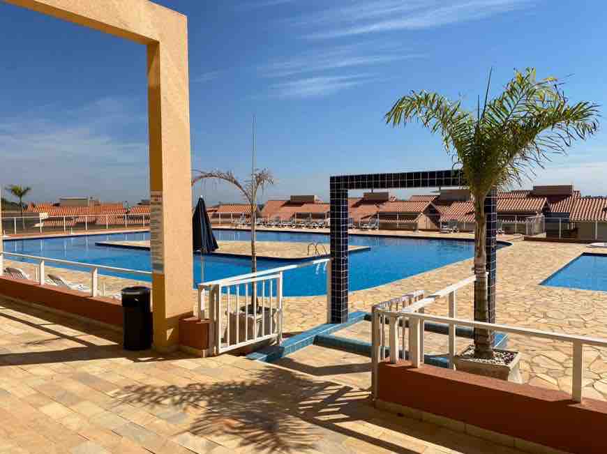 A wide view of the swimming pool area is presented, featuring two swimming pools flanked by palm trees. Surrounding the pools, lounge chairs are positioned on the stone deck, with umbrellas providing shade. The clear blue sky adds to the ambient atmosphere.