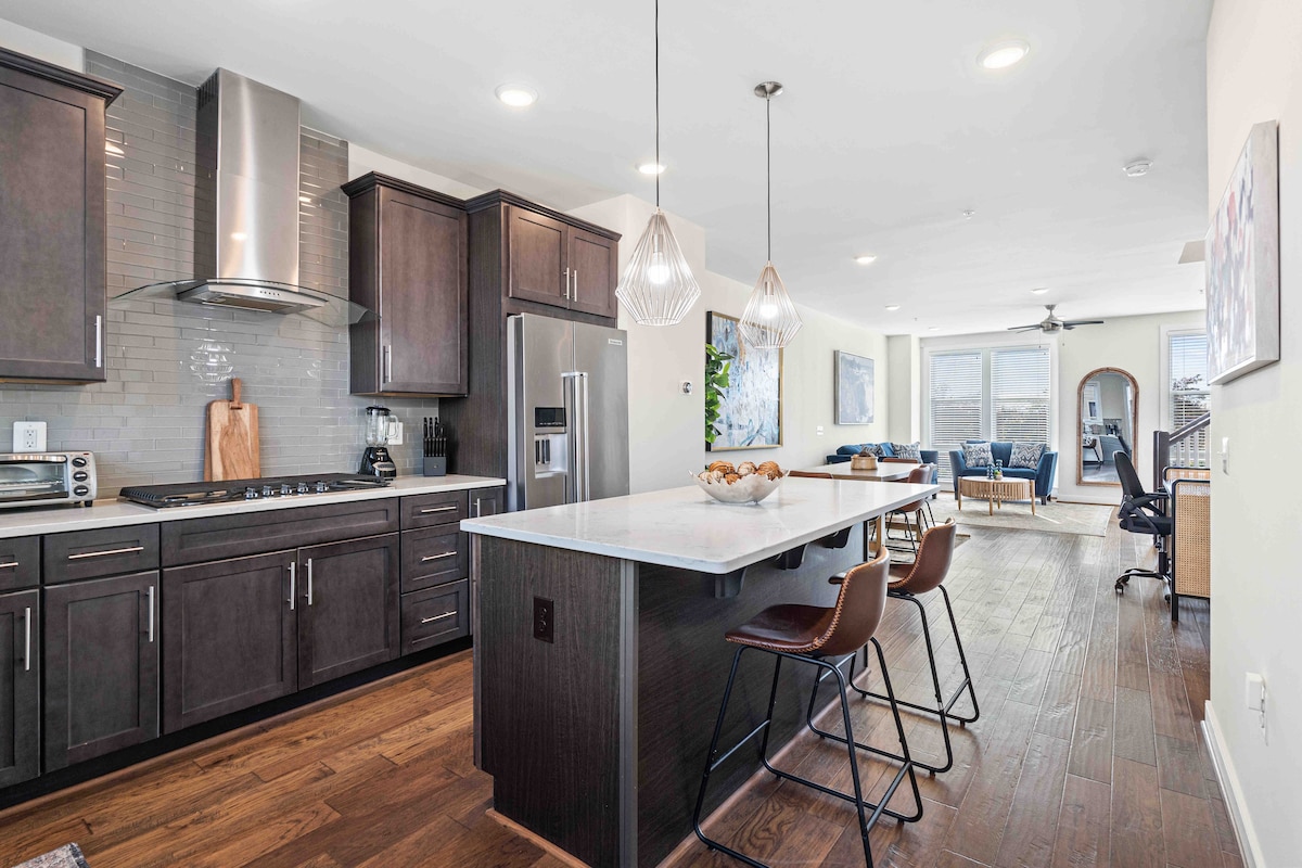A modern kitchen is highlighted with dark wood cabinetry and stainless steel appliances. A large island with seating offers ample space, while natural light fills the open living area visible in the background, featuring comfortable seating and large windows.