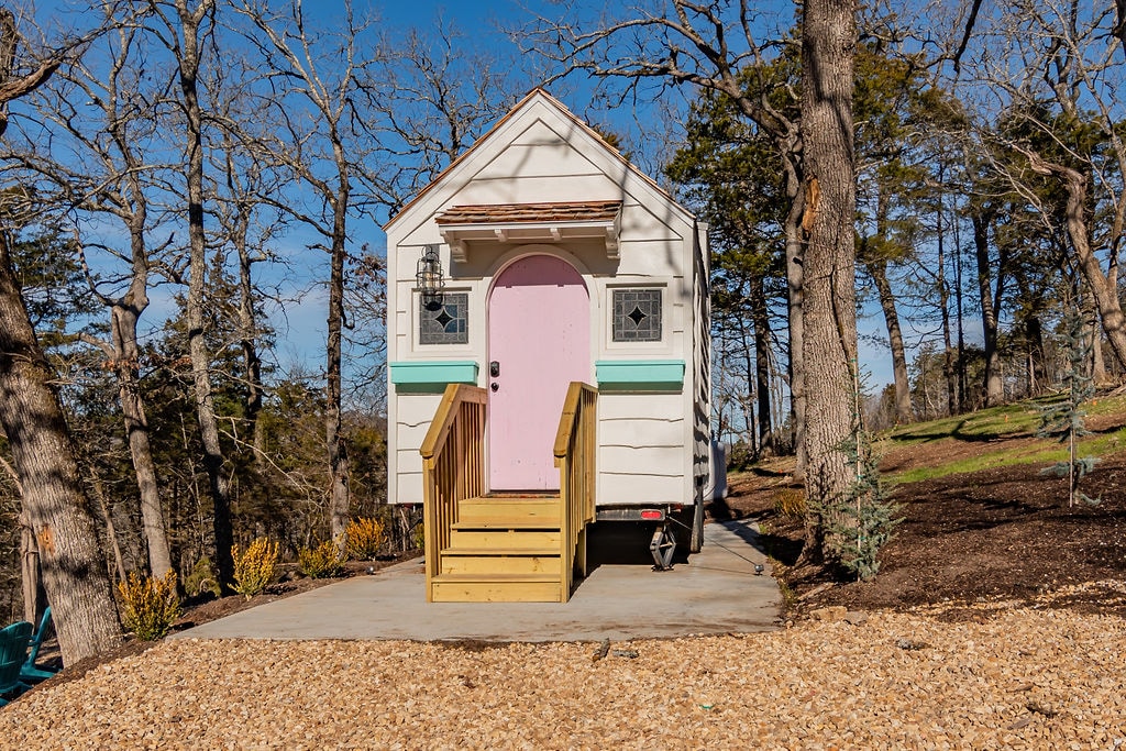 The tiny home is situated on a solid foundation, featuring a light exterior with a distinct pink door. Steps lead up to the entrance, surrounded by natural landscaping and a canopy of trees, providing a peaceful retreat in the woods.