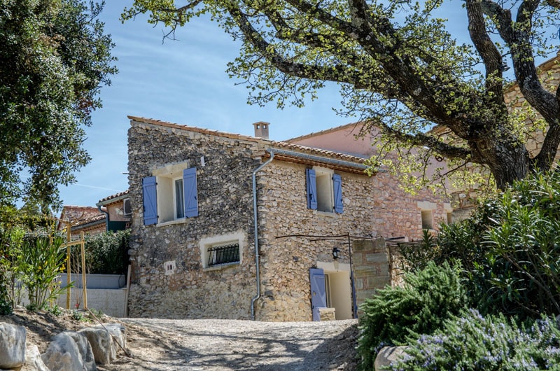 A charming stone house with blue shutters is situated among greenery in a rural setting. The building showcases its rustic character with a blend of stone and plaster. Surrounding vegetation adds to the natural ambiance, while a gravel pathway leads up to the entrance.