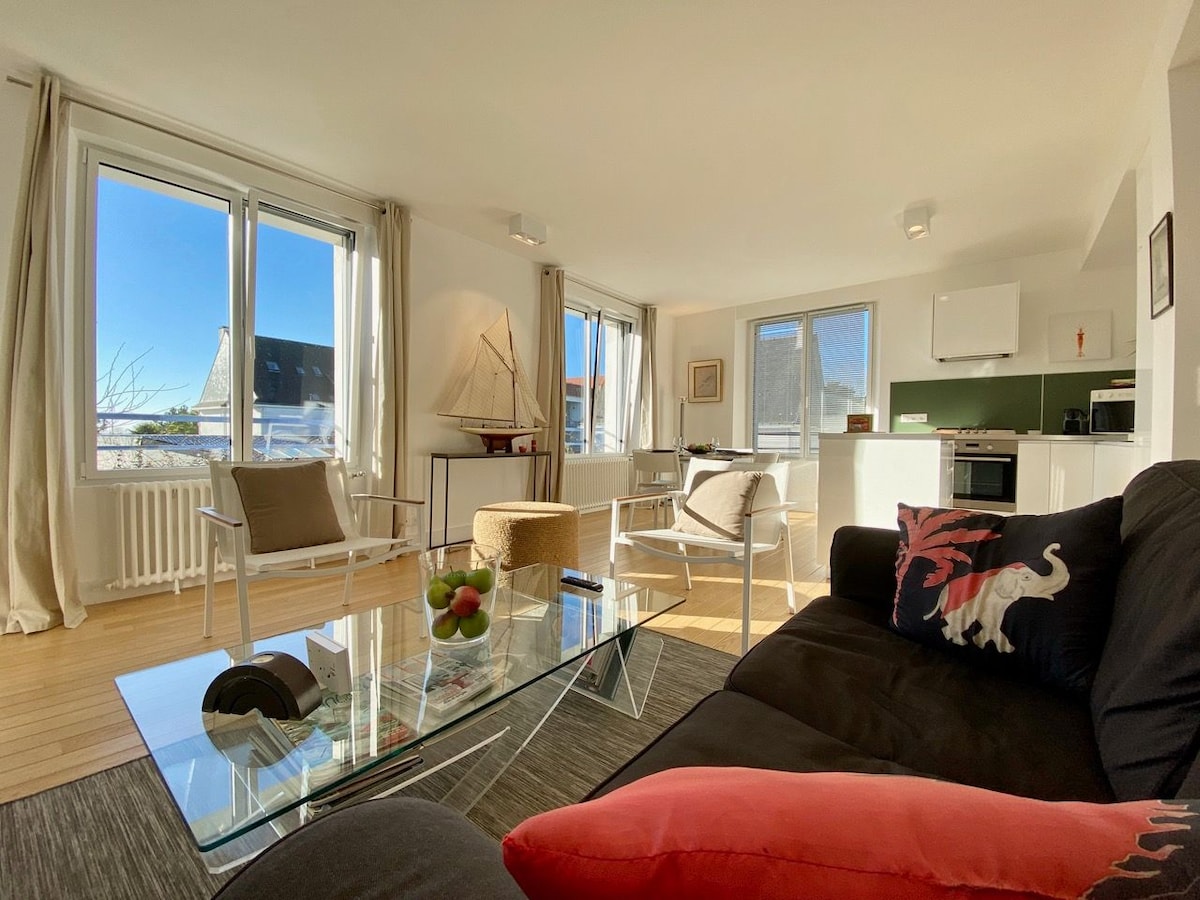 A bright living space is highlighted by large windows allowing natural light. Comfortable seating is arranged around a glass coffee table, complemented by decorative items. The open kitchen is visible in the background, equipped with modern appliances and sleek green accents.