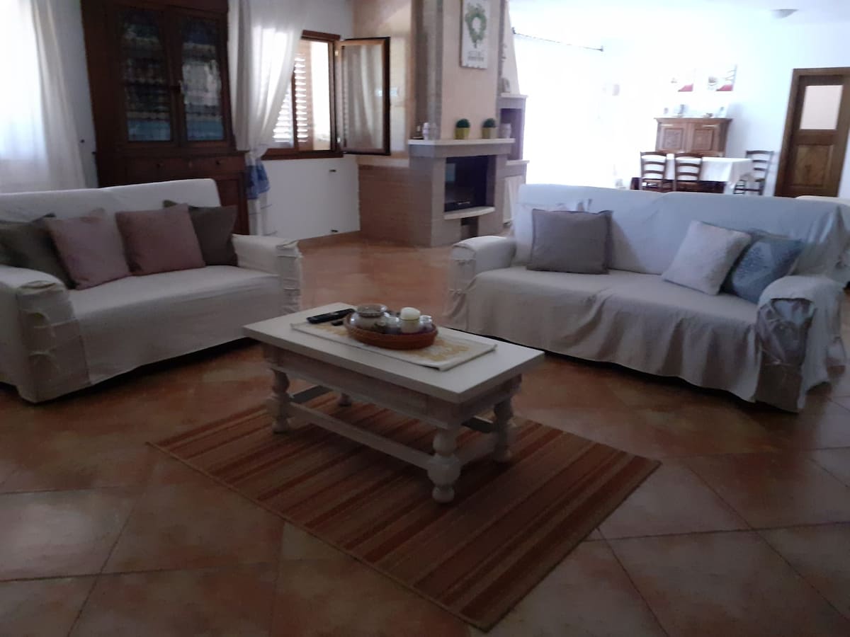 A living area is furnished with two white sofas, each adorned with decorative cushions. A wooden coffee table is placed at the center, holding a tray with small decorative items. Light enters through large windows, illuminating the tile floor and creating a cozy ambiance.