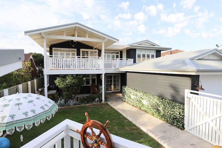 A two-story house with a white balcony and trim, featuring a nautical theme with a decorative ship wheel on the front porch. The house has a well-kept yard with a pathway leading to the entrance and ivy growing along the side of the building